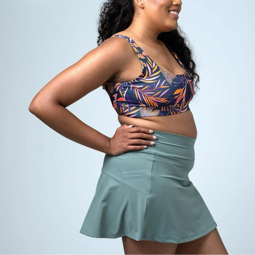 Woman modelling a sage green athletic skort with perforated side panels and a colourful printed sports bra.