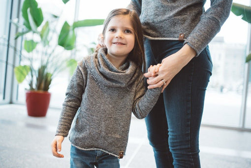 Young child in a grey knit top holding an adult’s hand in a bright indoor space.
