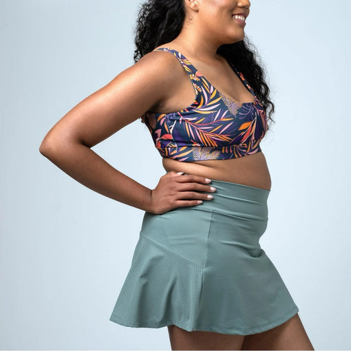 Woman modelling a sage green athletic skort with perforated side panels and a colourful printed sports bra.