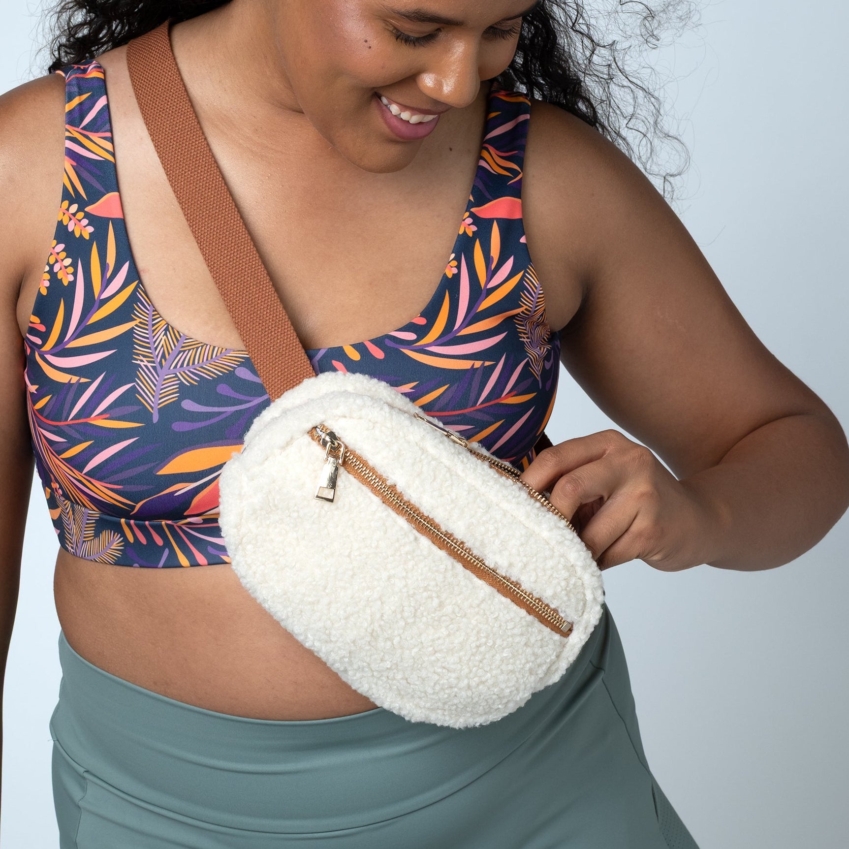 A woman wearing a colorful tropical print sports bra and a sage green skirt models a cream-colored fuzzy crossbody bag with a brown strap and zipper.