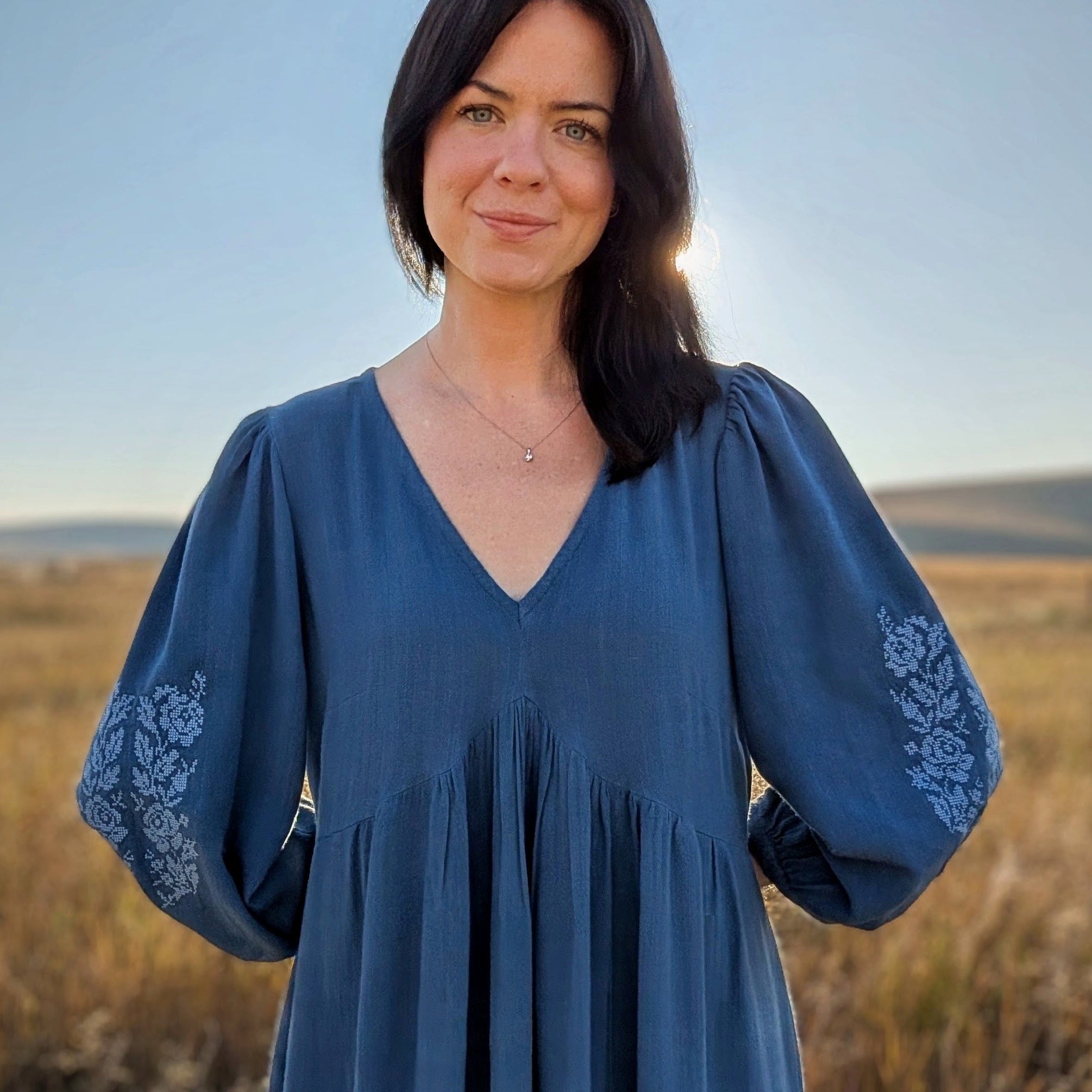 A woman with dark hair stands outdoors in a field wearing a blue long-sleeve dress with white floral embroidery on the sleeves. The sun is setting behind her, creating a warm glow, and the background shows dry grass and a clear blue sky.