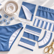 A styled flat-lay of sewing and crafting supplies, including a pair of blue and white bikini-style underwear, fabric pieces, striped trim, thread, clips, a bowl of elastic, and gold scissors on a white surface.