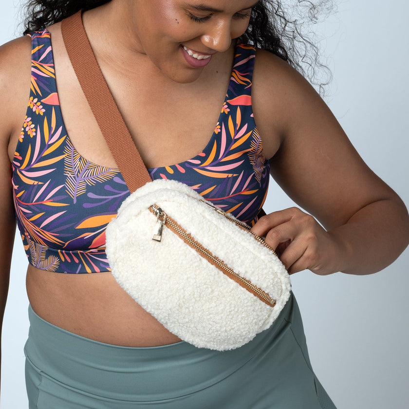 A woman wearing a colorful tropical print sports bra and a sage green skirt models a cream-colored fuzzy crossbody bag with a brown strap and zipper.
